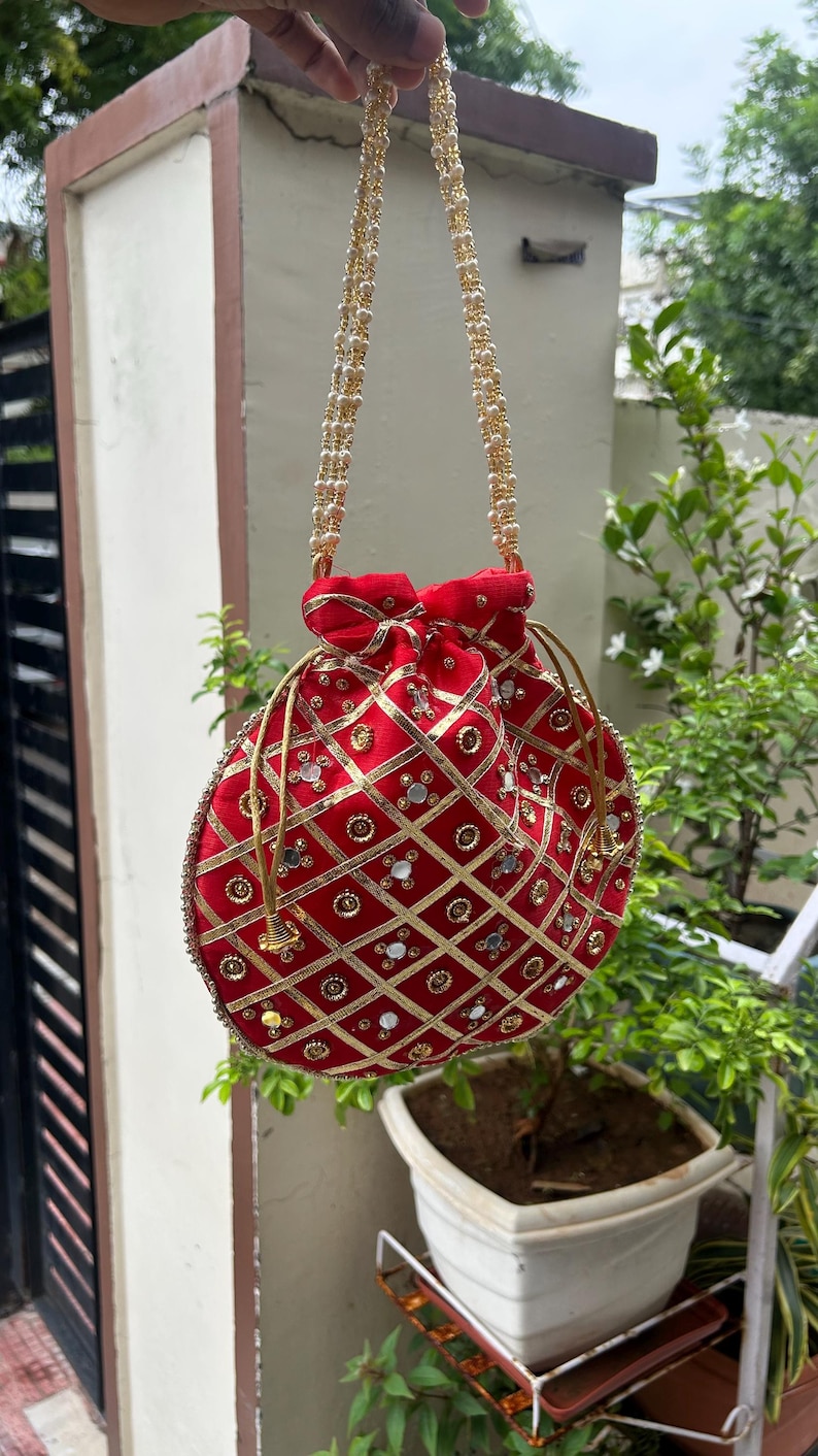 K&ouml;nnte beinhalten: Eine rote, runde Beuteltasche mit goldenen Verzierungen und einem goldperlenbesetzten Riemen. Die Tasche hat ein Steppmuster mit goldenen Zierleisten und kleinen, reflektierenden Akzenten. Die Tasche h&auml;ngt vor einem gr&uuml;nen, bl&auml;ttrigen Hintergrund.