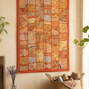 May include: A decorative patchwork wall hanging in shades of orange, gold, and cream. The rectangular textile has a solid orange border and is made up of various patterned fabrics. It is displayed on a light-coloured wall, adding a touch of warmth.