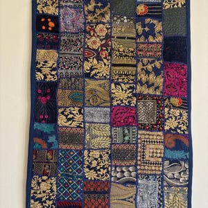 Puede incluir: Un tapiz rectangular con un diseño de retazos de varios cuadrados de tela. El tejido presenta una base azul oscuro con intrincados bordados en hilos dorados, rosas, rojos y verdes, mostrando patrones florales, geométricos y de cachemira.