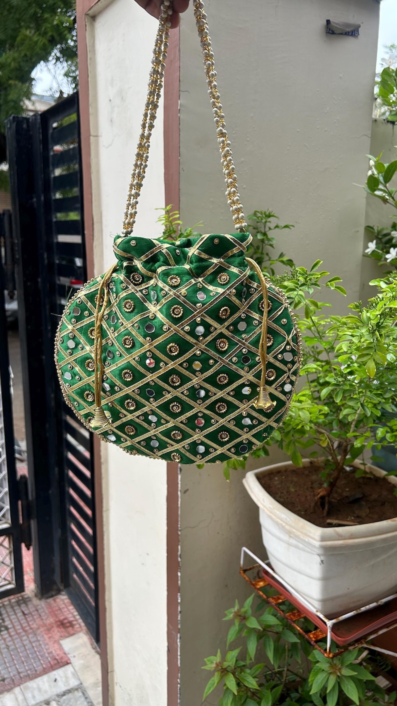 K&ouml;nnte beinhalten: Eine gr&uuml;ne, runde Beuteltasche mit goldenen Verzierungen und Spiegelakzenten. Die Tasche hat ein Rautenmuster mit goldenem Besatz und einen goldfarbenen Perlenhenkel. Die Tasche h&auml;ngt vor einem verschwommenen Hintergrund.