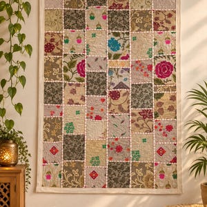 Op de afbeelding: Een rechthoekige wandhanger met een patchworkontwerp van verschillende stoffen vierkanten. Het textiel toont een mix van bloemen-, paisley- en geometrische patronen in tinten groen, beige, roze en blauw, verbonden door witte decoratieve stiksels.