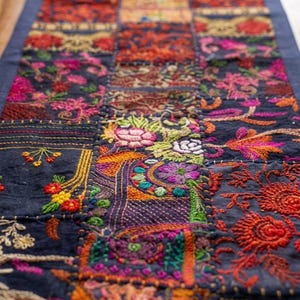 Pode incluir: Um caminho de mesa retangular com um design de retalhos de quadrados de tecido azul escuro. Cada quadrado &eacute; decorado com bordados florais e geom&eacute;tricos complexos e multicoloridos em tons de vermelho, laranja, rosa, roxo, verde e amarelo.