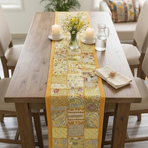 Op de afbeelding: Een houten eettafel met een gele patchwork tafelloper met paisley- en bloemmotieven. Twee brandende kaarsen, een vaas met bloemen en een waterkan staan op de tafel. Beige stoelen omringen de tafel. De tafel is gedekt voor een maaltijd.