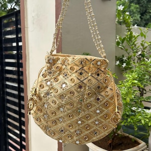 K&ouml;nnte beinhalten: Eine goldfarbene, runde Handtasche mit Kordelzugverschluss und Perlenriemen. Die Tasche ist mit kleinen Spiegeln und goldfarbenen Zierelementen in Rautenmuster verziert. Eine Quaste h&auml;ngt an der Seite.