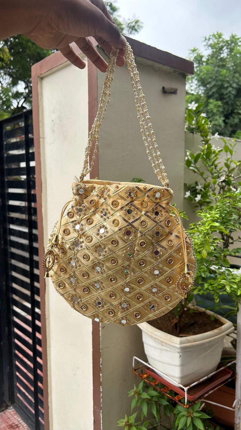 K&ouml;nnte beinhalten: Eine goldfarbene, runde Handtasche mit Kordelzugverschluss und Perlenriemen. Die Tasche ist mit kleinen Spiegeln und goldfarbenen Zierelementen in Rautenmuster verziert. Eine Quaste h&auml;ngt an der Seite.