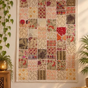 May include: A rectangular wall hanging featuring a patchwork design of various fabric squares. Each square displays unique embroidery, floral patterns, and textures in shades of beige, red, pink, and brown, all joined by visible white stitching.