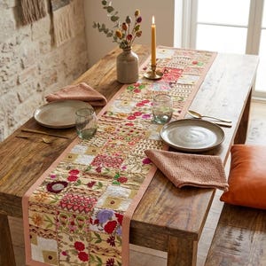 Peut inclure: Un chemin de table rectangulaire présentant un motif patchwork de broderies florales dans des tons de rouge, rose, jaune et vert sur une base en tissu beige clair, fini avec une bordure unie couleur pêche sur les bords longs.