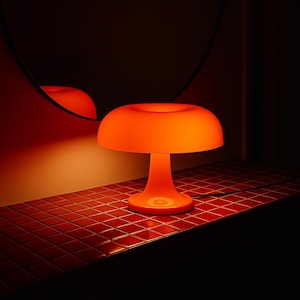 Puede incluir: Una lámpara de mesa naranja con forma de hongo, con base redonda y pantalla ancha y curvada. La lámpara es de material sólido y está sobre una superficie de baldosas rojas. La lámpara está enchufada.