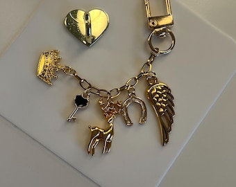 Op de afbeelding: Goudkleurige sleutelhanger met diverse bedels, waaronder een kroon, sleutel, hert, hoefijzer en vleugel. Er is ook een gouden hartvormige haak aanwezig. De bedels zijn bevestigd aan een ketting en een sluiting. Witte achtergrond.