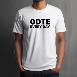 Puede incluir: Camiseta blanca con el texto negro "ODTE EVERY DAY" impreso en la parte delantera. La camiseta es de manga corta y tiene cuello redondo. La persona que lleva la camiseta tiene las manos en los bolsillos.