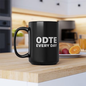Puede incluir: Una taza de cerámica negra con un asa grande. La taza presenta el texto blanco "ODTE EVERY DAY" en la parte delantera. La taza está sobre una superficie de madera, con un fondo borroso.