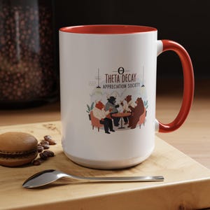 May include: White ceramic mug with a red interior and handle. The mug displays the text "THETA DECAY APPRECIATION SOCIETY" and an illustration of animal figures in a cafe. A macaron and spoon are on the wooden surface.