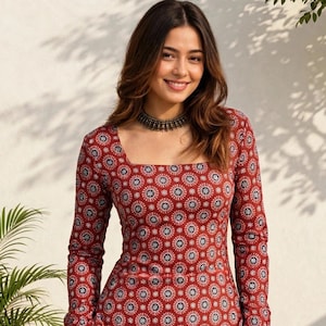 May include: A long-sleeved tunic featuring a square neckline and a repeating circular geometric pattern in shades of deep red, white, and black. The garment has a fitted bodice, straight sleeves, and side pockets, paired with plain white trousers.