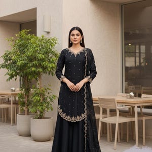 Op de afbeelding: Een zwart ensemble met een top met lange mouwen met gouden borduursels en kleine details, gecombineerd met een bijpassende rok. Een doorschijnende, bedrukte sjaal is aan de zijkant gedrapeerd. De tekst "001fashionforward" is zichtbaar.