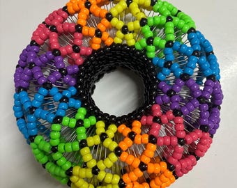 Peut inclure: Un objet circulaire perlé coloré avec un centre noir. Les perles sont disposées en arc-en-ciel, avec des sections rouges, oranges, jaunes, vertes, bleues et violettes. Les perles sont reliées par du fil d'argent.
