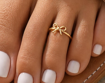 Könnte beinhalten: Ein Zehenring aus goldfarbenem Metall mit einem kleinen, zierlichen Schleifendesign in der Mitte. Der Ring wird am zweiten Zeh getragen, der mit weißem Nagellack lackiert ist, und der Fuß ruht auf einer beigen Ledersandale.