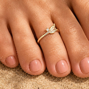 Op de afbeelding: Een goudkleurige teenring met een delicaat bladmotief en een klein, helder edelsteenaccent. De ring is ontworpen met een slanke, gepolijste band die om de teen past en een botanisch geïnspireerde esthetiek met een metallic afwerking laat zien.