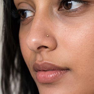 May include: A small, round, gold-coloured nose stud featuring a smooth, polished surface. The jewellery is positioned on the side of the nostril, showcasing a minimalist design that sits flush against the skin with a subtle metallic lustre.