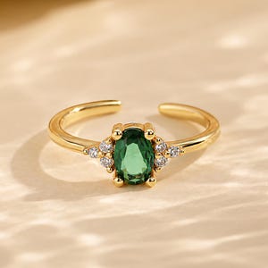 Puede incluir: Un anillo abierto de tono dorado con una piedra preciosa verde de corte ovalado en el centro, flanqueada por tres pequeñas piedras transparentes a cada lado. La banda es pulida y lisa, con una abertura en la parte posterior del anillo.