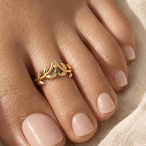 Könnte beinhalten: Ein goldener Zehenring mit einem zarten, von Blättern inspirierten Banddesign, das sich zu einem zentralen Punkt wölbt. Der Ring ist mit einem einzelnen, kleinen, birnenförmigen grünen Edelstein an der Spitze des Blattmotivs verziert.