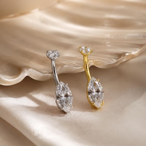 May include: Two curved barbell belly button rings, one in silver-tone metal and one in gold-tone metal. Each piece features a small round clear gemstone at the top and a larger marquise-cut clear gemstone at the bottom, held by metal prongs.