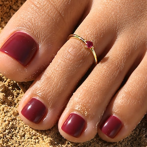 May include: A thin gold toe ring featuring a small, round, faceted red gemstone set in the center. The ring is worn on the second toe, which is painted with a glossy, dark burgundy nail polish, matching the polish on the other visible toes.