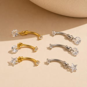 May include: A collection of six curved barbell body jewelry pieces. Three are gold-toned and three are silver-toned. Each features a clear, round, or star-shaped cubic zirconia charm dangling from one end, with a matching stone at the other end.