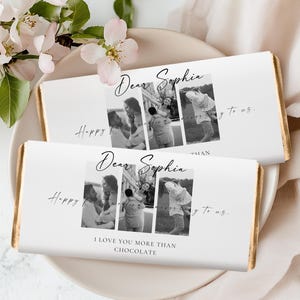 Könnte beinhalten: Zwei Schokoriegel mit weißen Verpackungen werden auf einem weißen Teller präsentiert. Jede Verpackung zeigt eine Fotocollage, die Worte "Dear Sophia" und den Satz "I love you more than chocolate". Die Riegel haben goldfarbene Folienkanten.
