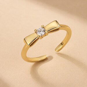 May include: A gold-toned open ring featuring a central bow-shaped design element. The center of the bow is accented with a single, clear, round-cut gemstone. The band is smooth and polished, with an adjustable opening at the back of the ring.