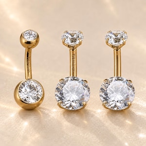 May include: Three gold-toned titanium navel rings featuring clear, round-cut faceted stones. The left piece has a bezel-set top and bottom stone, while the two right pieces feature prong-set stones. Each curved barbell is engraved with the word Titanium.