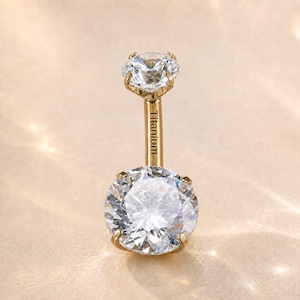May include: A gold-toned belly button ring featuring a large round clear gemstone at the base and a smaller round clear gemstone at the top. The straight post connecting the two stones is engraved with the word Titanium in black lettering.