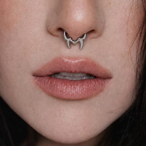 May include: A silver septum ring featuring a symmetrical, pointed design that resembles two downward-curving fangs or horns. The jewelry has a polished, metallic finish and is positioned securely within the nasal septum, centered above the upper lip.