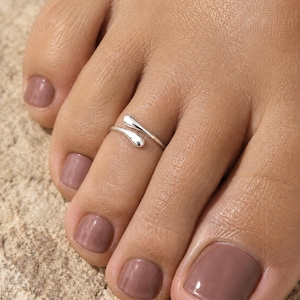 Può includere: Un anello da dito del piede aperto color argento con due estremità lisce e arrotondate a forma di goccia che si sovrappongono leggermente. La fascia in metallo ha una finitura lucida e riflettente ed è indossata sul secondo dito del piede.