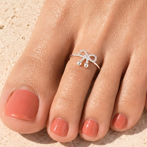 Puede incluir: Un anillo para el dedo del pie de tono plateado con un delicado diseño de lazo y dos pequeñas esferas colgantes. El anillo se lleva en el segundo dedo, que tiene esmalte de uñas color coral. La banda metálica es fina, pulida y abierta.