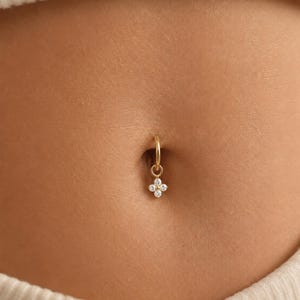 Könnte beinhalten: Ein goldfarbener Bauchnabelpiercing mit einem kreisförmigen Ring und einem hängenden Anhänger in Form einer vierblättrigen Blume. Der Blumenanhänger ist mit vier kleinen, klaren, rund geschliffenen Steinen besetzt, die das Licht reflektieren.