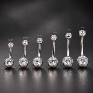 May include: Six curved barbell navel rings made of silver-toned titanium, each featuring a clear round-cut gemstone at both ends. The lengths are labelled as 6mm, 8mm, 10mm, 12mm, 14mm, and 16mm. Each barbell is engraved with the word Titanium.