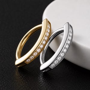May include: Two circular hoop rings, one in a polished gold tone and one in a polished silver tone. Each ring features a flat, wide front section embedded with a row of small, clear, round-cut stones that catch the light, creating a sparkling effect.