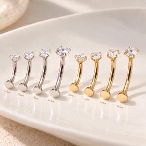 May include: A collection of eight curved barbell body jewelry pieces with clear round gemstones. Four are silver-toned and four are gold-toned, with lengths labeled 10mm, 12mm, 14mm, and 16mm. Each piece features a flat circular base and a prong-set stone.