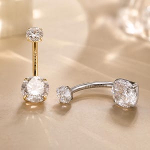 May include: Two navel rings featuring clear round-cut gemstones set in metal. One ring is gold-toned with a vertical post, while the other is silver-toned with a curved post. Both posts are clearly engraved with the word Titanium for identification.