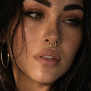 Puede incluir: Un anillo de septum dorado que presenta una serie de eslabones circulares texturizados e interconectados que forman un diseño decorativo similar a una cadena. La joya está colocada en el centro de la nariz, mostrando un acabado metálico.