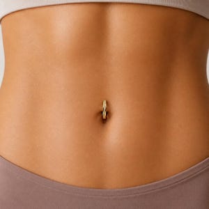 May include: A gold-toned circular navel ring featuring a row of small, clear, sparkling stones embedded along the front surface. The jewellery is positioned securely within the belly button, showcasing a polished metallic finish and a classic hoop design.
