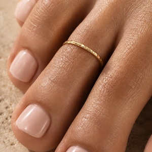 Puede incluir: Un anillo para el dedo del pie fino de color dorado con una superficie texturizada y martillada. La banda es circular y se coloca alrededor del segundo dedo, mostrando un acabado metálico reflectante con pequeñas facetas.