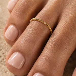 Op de afbeelding: Een dunne, goudkleurige teenring met een delicaat, getextureerd, kraalachtig oppervlak. De ring wordt gedragen om de tweede teen, die is voorzien van lichtgekleurde nagellak. Het ontwerp is eenvoudig, cirkelvormig en metaalachtig van aard.