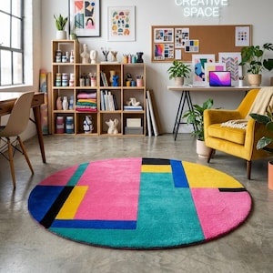 Könnte beinhalten: Ein farbenfroher, runder Teppich mit geometrischem Design. Der Teppich zeigt große Blöcke in Pink, Türkis, Gelb, Blau und Schwarz. Der Teppich befindet sich in einem Raum mit einem Schreibtisch, einem Stuhl und einem gelben Sessel.