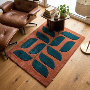 May include: A rectangular rug with a leaf pattern in a living room setting. The rug has a terracotta base with teal leaf shapes. The rug is placed on a wooden floor, and the scene includes a brown leather chair and a small wooden table. The rug measures approximately 150cm x 90cm.