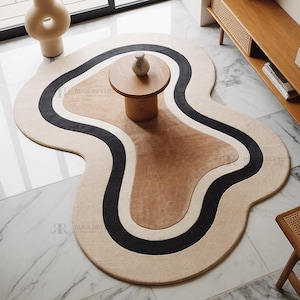Könnte beinhalten: Ein abstrakt geformter Teppich mit einem beige-, braun-, schwarz- und weißen Design. Der Teppich hat eine wellenförmige, organische Form und liegt auf einem Marmorboden. Ein kleiner, runder Holztisch steht auf dem Teppich.