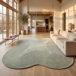 May include: A large, sage green area rug with an irregular, organic shape and a textured surface featuring concentric, raised circular lines that follow the perimeter of the rug, creating a ripple-like pattern across the entire floor covering.