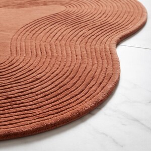 May include: A terracotta-colored rug with an irregular, organic shape and a textured surface featuring raised, concentric wavy lines that follow the perimeter of the piece, creating a rhythmic pattern across the entire top of the floor covering.