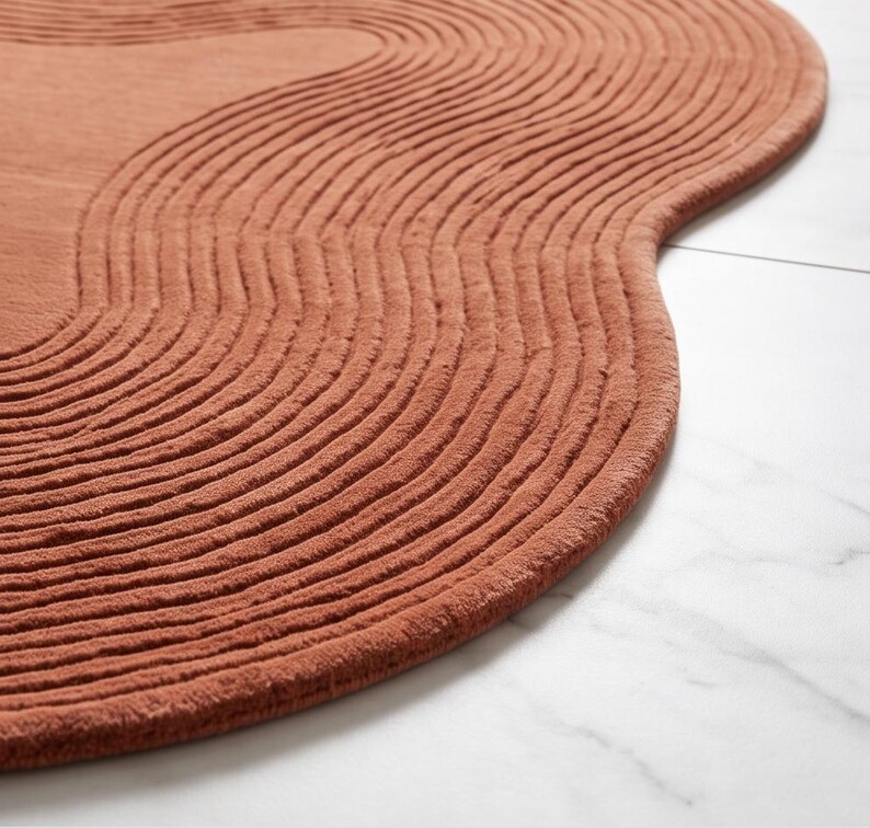 May include: A terracotta-colored rug with an irregular, organic shape and a textured surface featuring raised, concentric wavy lines that follow the perimeter of the piece, creating a rhythmic pattern across the entire top of the floor covering.