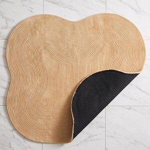 May include: An irregular, organic-shaped rug featuring a light tan, textured surface with concentric, wavy line patterns. The rug is partially folded over to reveal a solid black, circular-patterned underside, contrasting with the tan top.